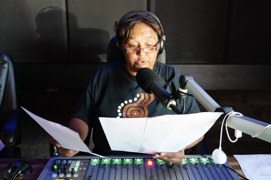 Pam Reilley wearing headphones and speaking into a microphone while reading from piece of paper, radio mixer in foreground
