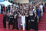 82 women stand on red-carpeted stairs in a block formation, with serious expressions, with Cannes signs in the background