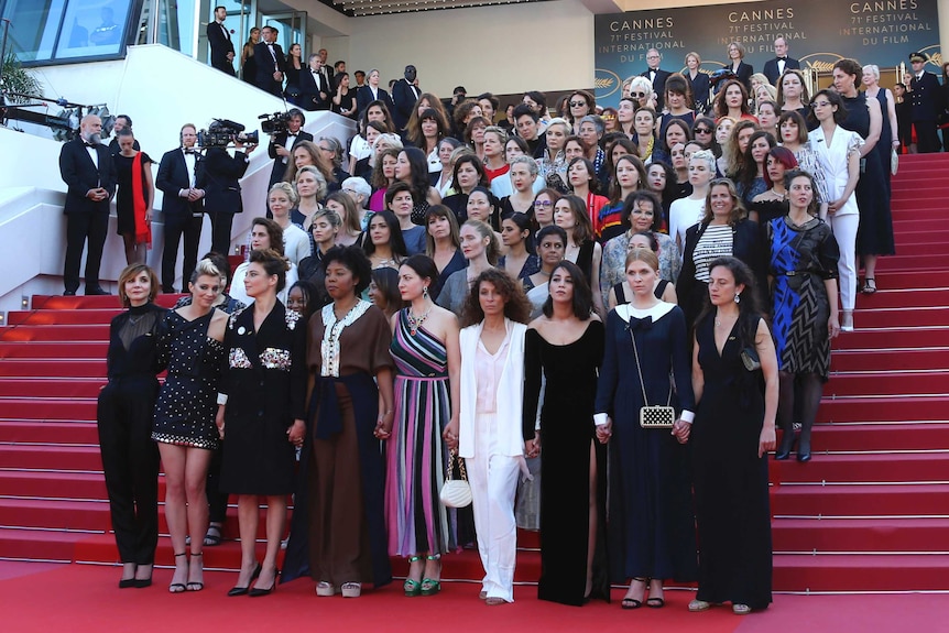82 women stand on red-carpeted stairs in a block formation, with serious expressions, with Cannes signs in the background