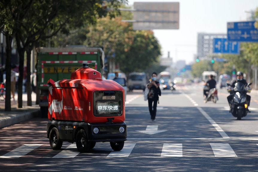 a red autonomous delivery vehicle travels across a pedestrian crossing during the day with motobikes and pedastrians nearby