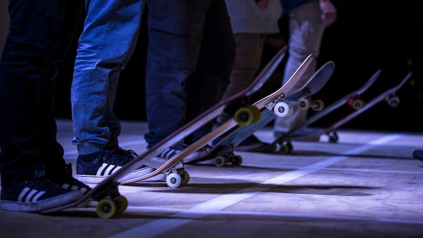 Five skateboarders seen from the waist down line up with a foot on their skateboards.