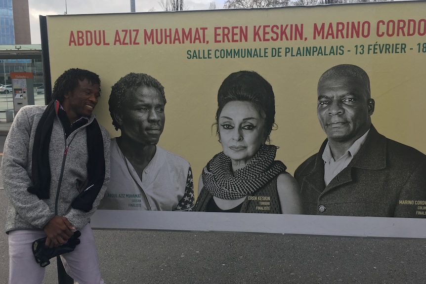 Man leans against a billboard smiling. He is featured on the billboard along with two other people.