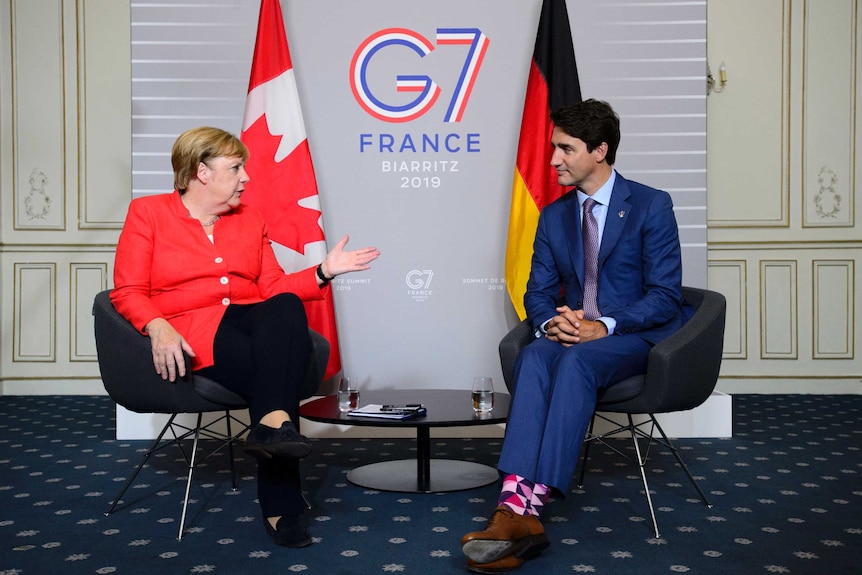 Merkel sits in front of Canadian flag while Trudeau sits in front of German flag.