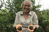 Dr Wendy Seabrook smiling at the camera with a weedy orchard behind her and soil in her hands.