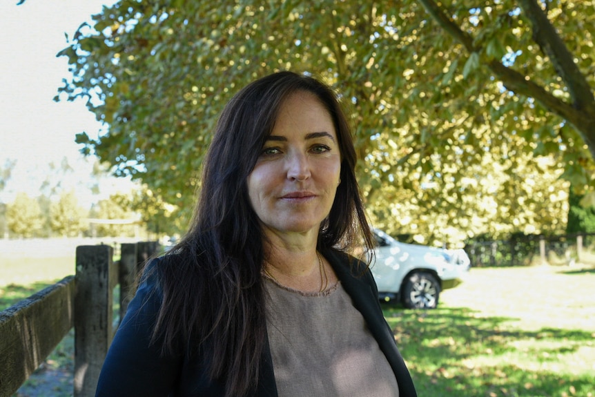 A woman with long dark brown hair stands in front of a wooden fence.