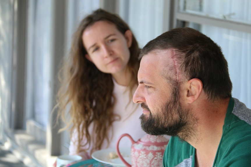 A couple sits on their balcony. The male is in the foreground and a large scar can be seen on the side of his head