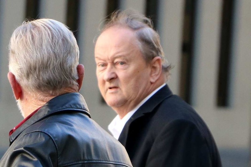 Two men wearing black jackets standing on a street, with one looking towards the camera.