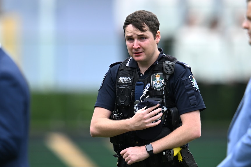 a queensland police officer in uniform holds their cap to their chest and looks upset