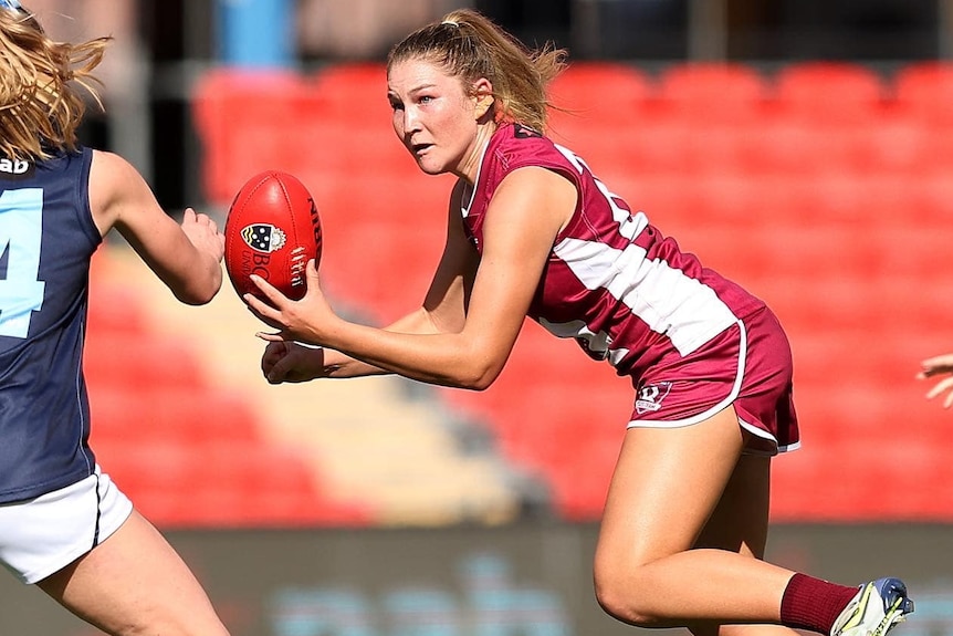 A young woman in a maroon AFL uniform catching a ball during a game.