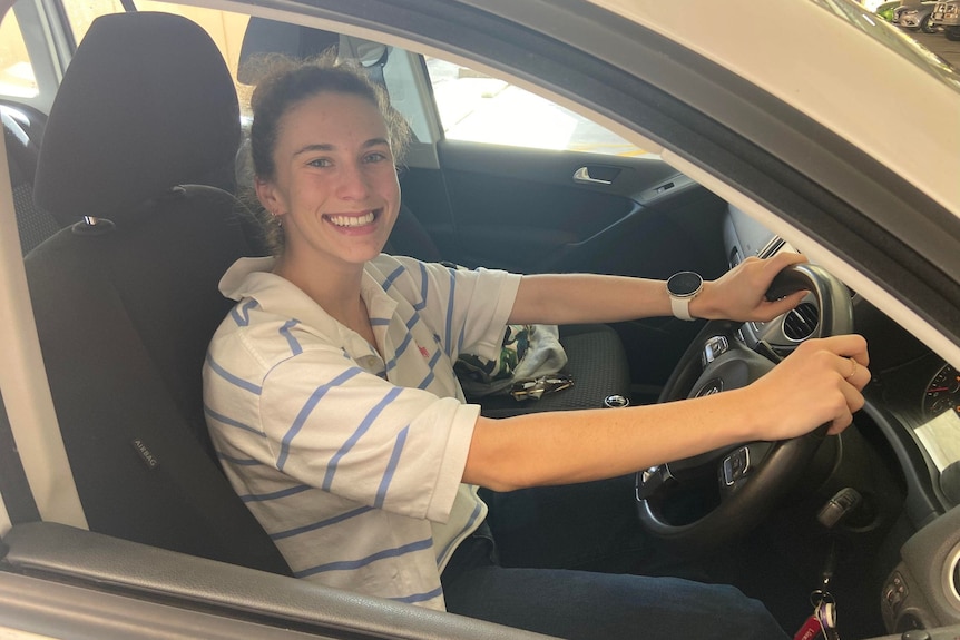 A woman in a car smiling at the camera with her hands on the steering wheel. 