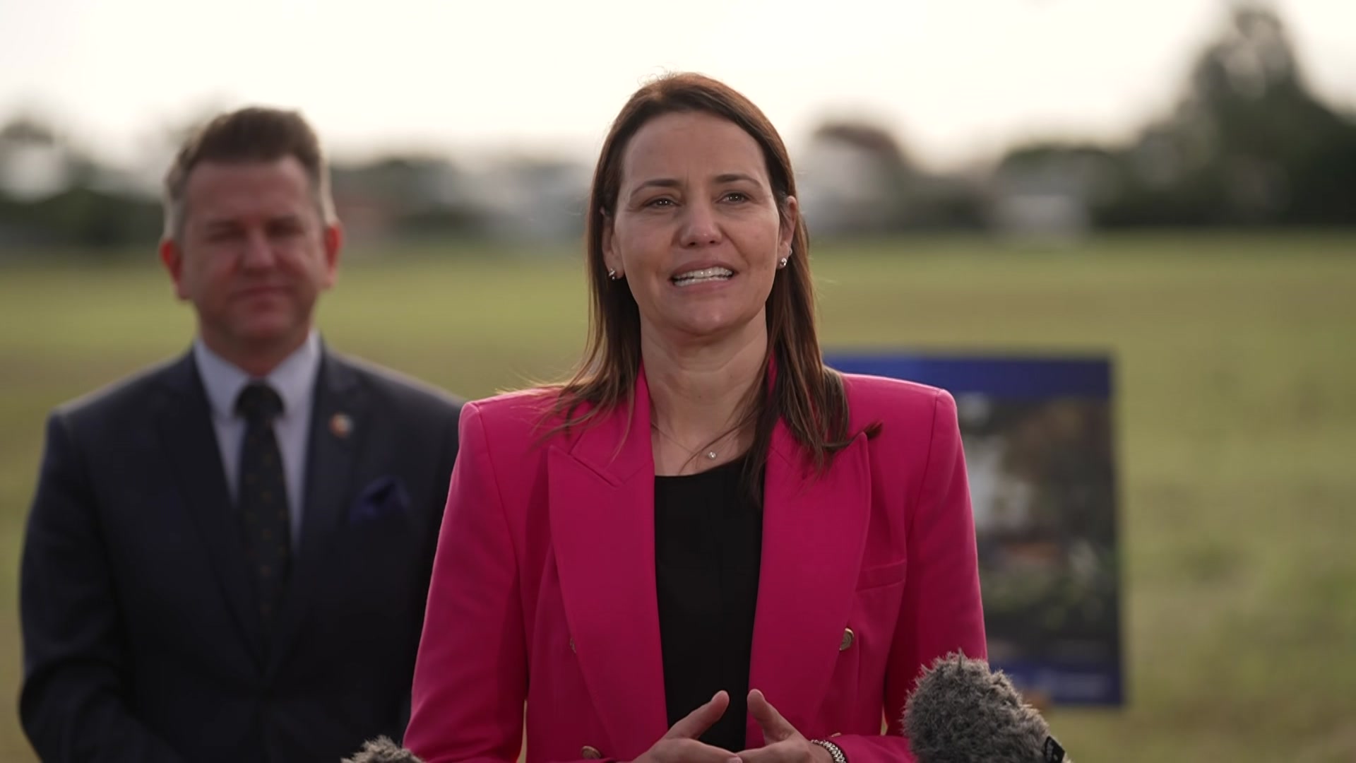 A woman in a pink blazer speaking into microphone with man in suit behind her.