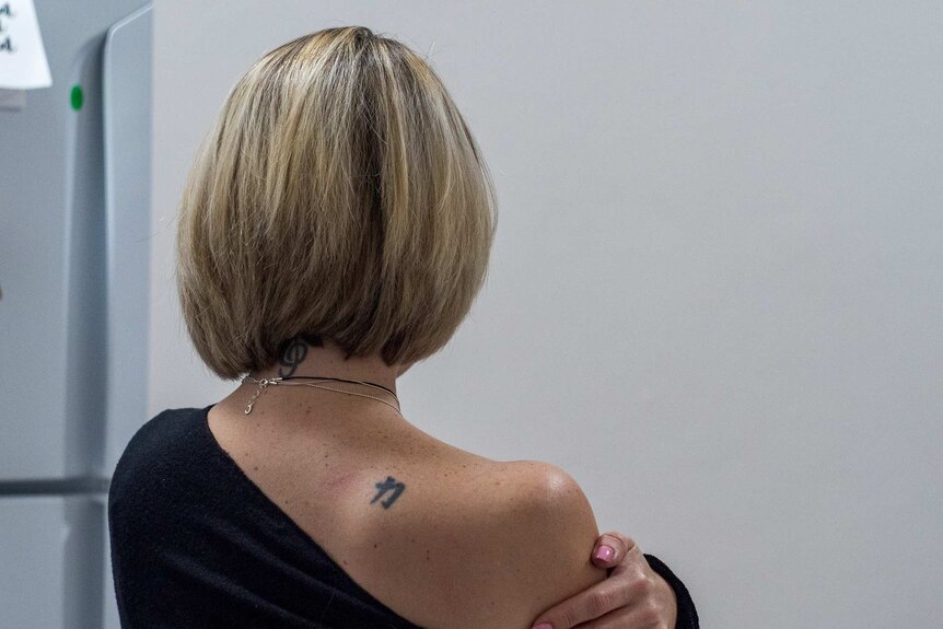 A photo of a woman with her back to camera, she is blonde and pulling down the shoulder of her top to reveal a tattoo.