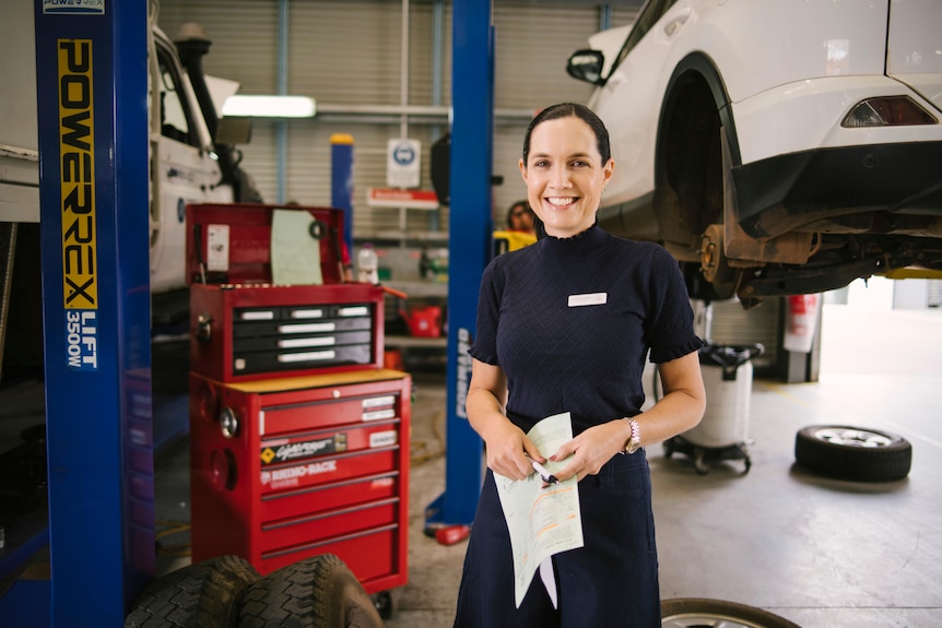 A woman in a blue shirt and dress pants stands in front of a mechanics workshop