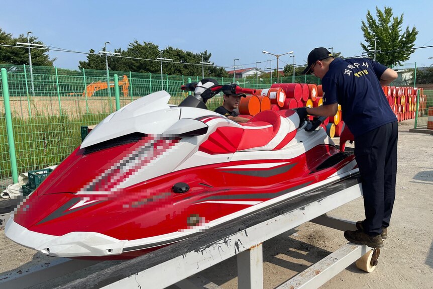 A South Korean coastguard officer inspects a red and white jet ski that sits on a small trailer behind a green fence.