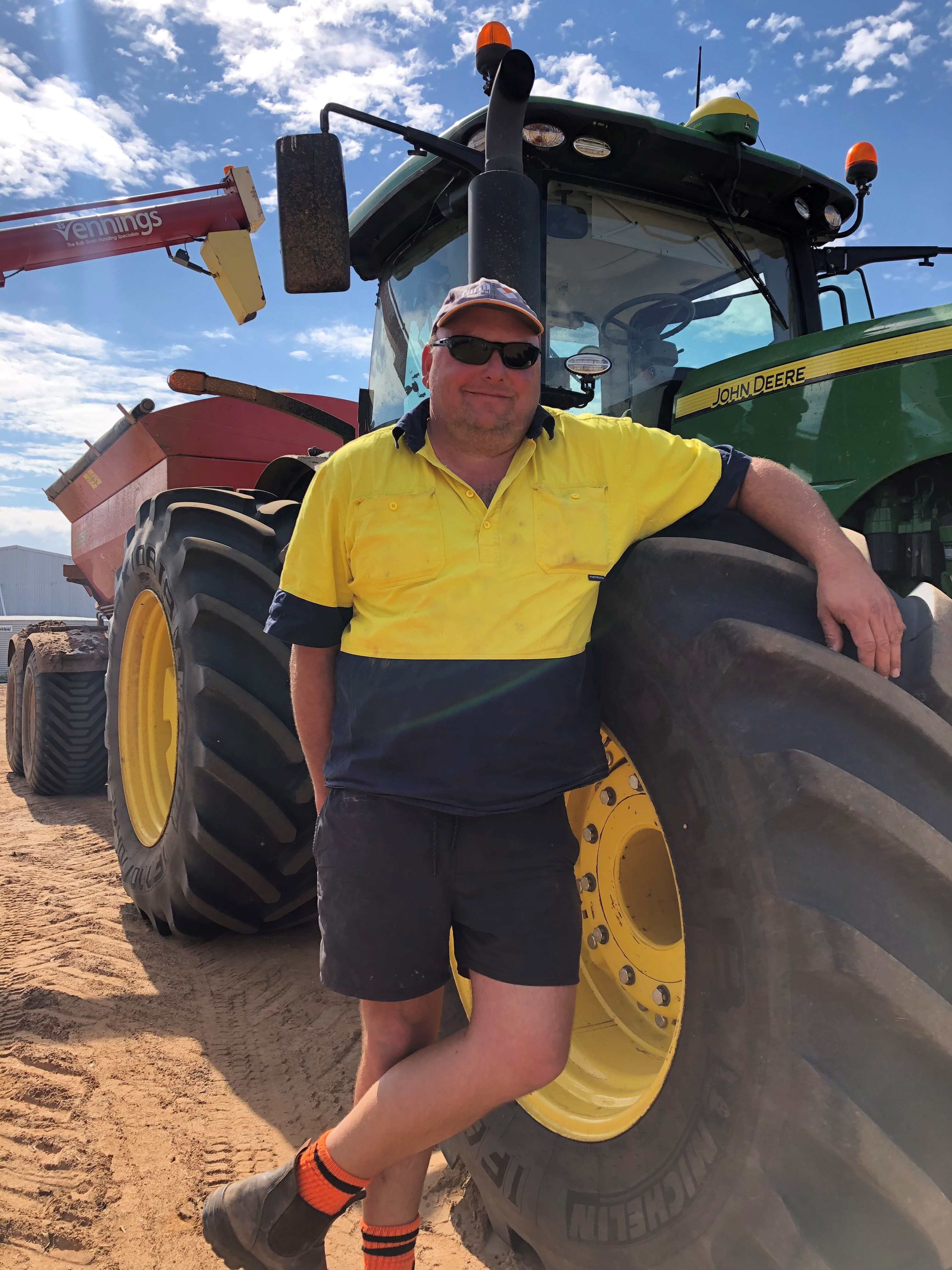 Man in yellow hi-vis shirt and shorts, wearing a light-coloured cap, leans against tractor wheel