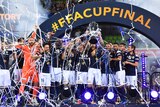 Melbourne Victory players in dark blue hold a trophy above their heads on stadium celebration platform with streamers and smoke.