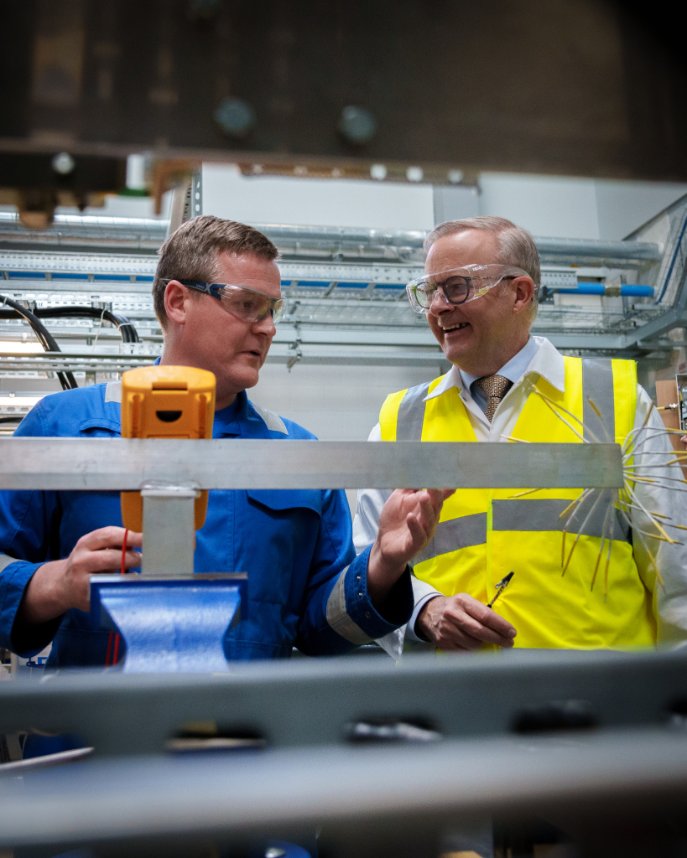 Albanese, in goggles and high vis, speaks to a man in blue coveralls. Obstructing the shot is a silver shelving unit
