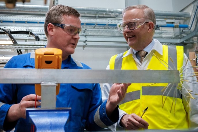 Albanese, in goggles and high vis, speaks to a man in blue coveralls. Obstructing the shot is a silver shelving unit