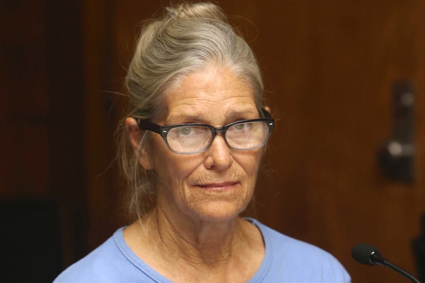 Leslie Van Houten looks to the camera with piercing expression. She wears a lilac top, her grey hair is up and has glasses on.