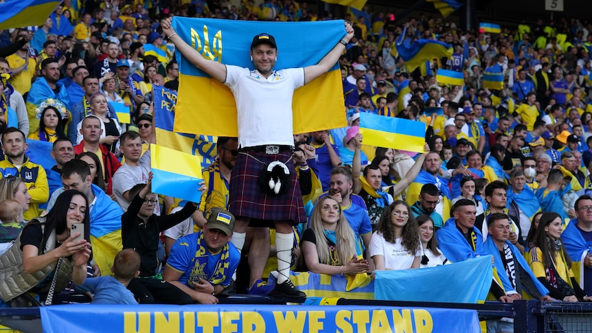 A man in a kilt holds a Ukraine flag above his head