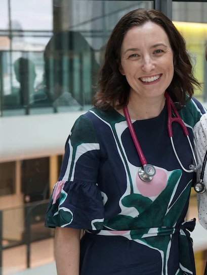 A woman wearing a stethascope and floral dress smiles at camera