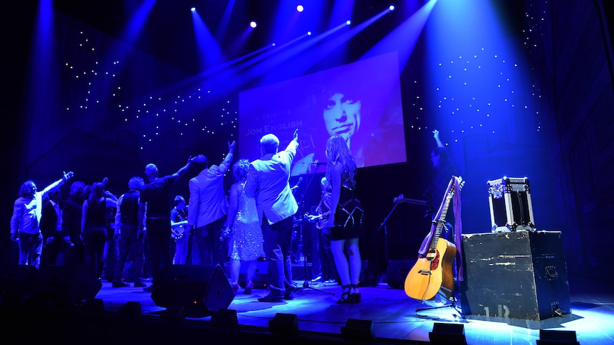 Friends and colleagues on stage point towards a photo of Jon English in tribute.