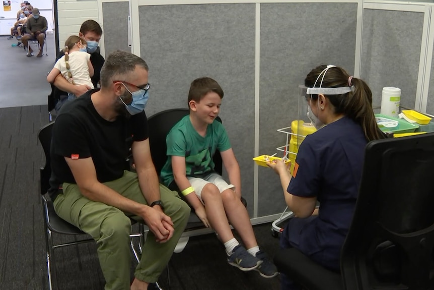 A man and a boy sit in a cubicle with a woman wearing a face shield