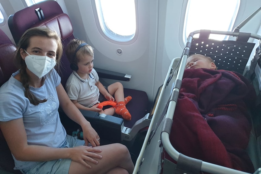 A woman with a mask on sitting on a plane next to a young boy with headphones with a baby sleeping in a travel bassinet infront