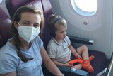 A woman with a mask on sitting on a plane next to a young boy with headphones with a baby sleeping in a travel bassinet infront