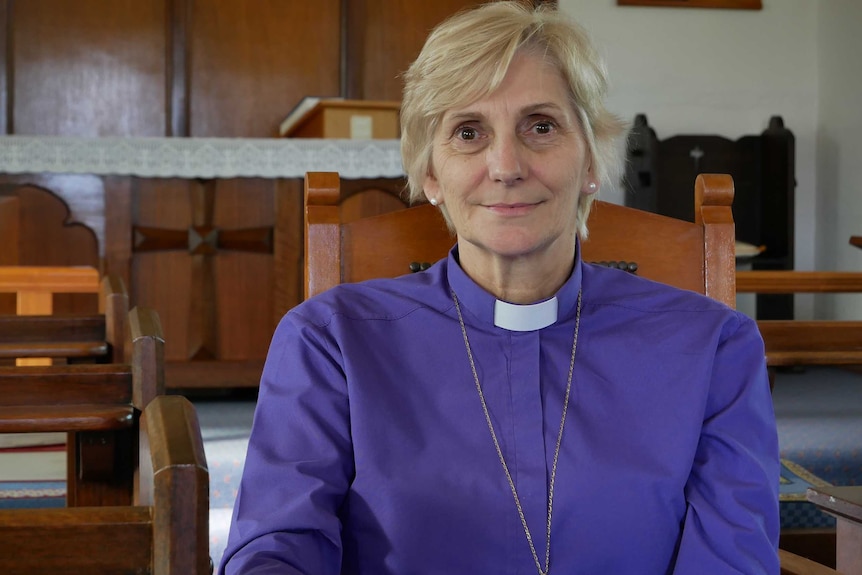 A female religious archbishop sits in a wooden chair inside a small rural church