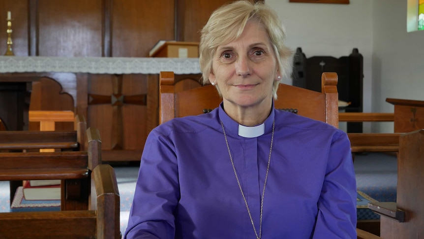 A female religious archbishop sits in a wooden chair inside a small rural church