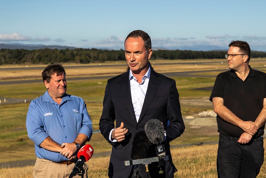 A man wearing a black suit stands behind a microphone speaking to media.