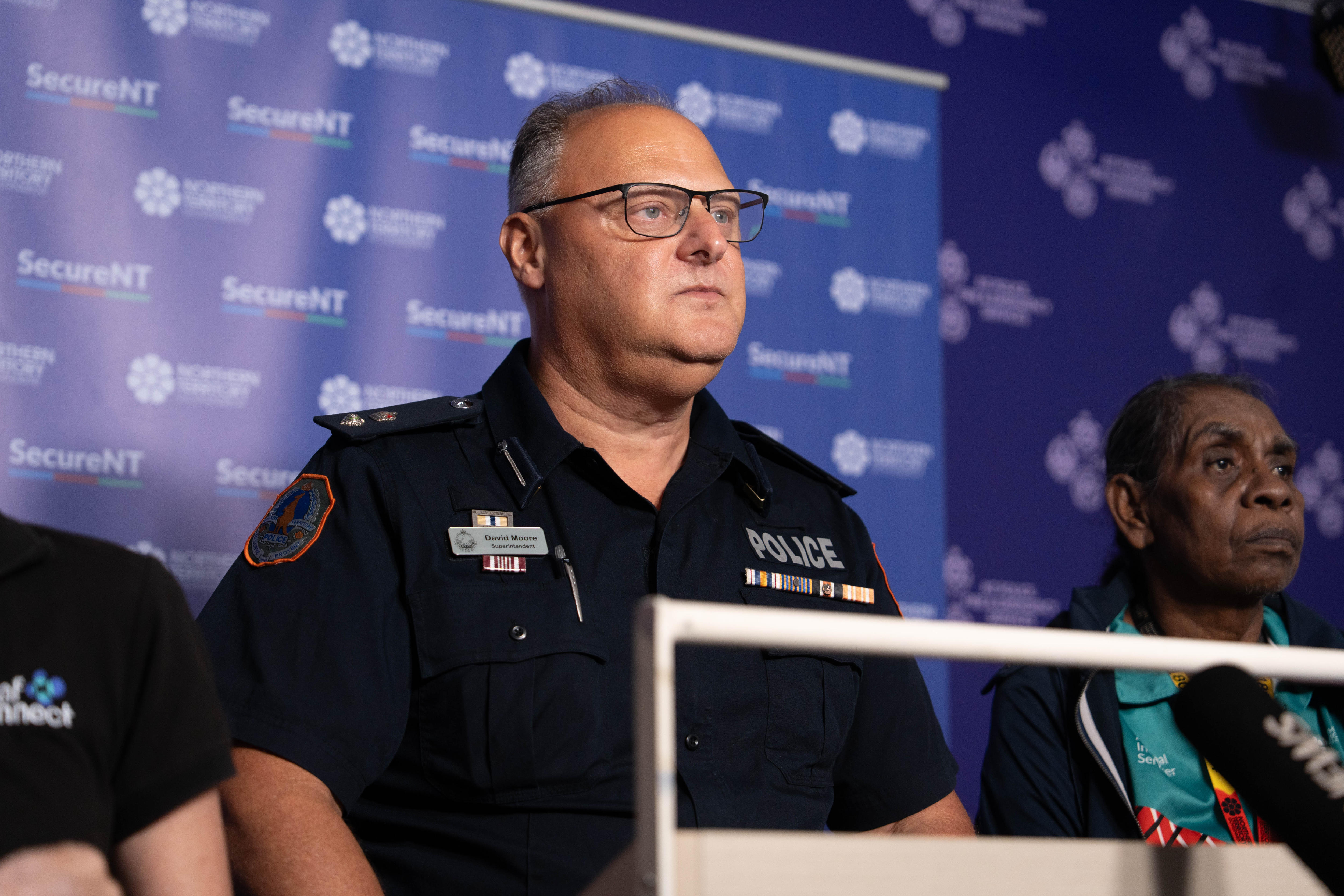 Hombre blanco, uniforme de policía negro, parado frente a una pancarta morada, expresión facial seria, gafas puestas.