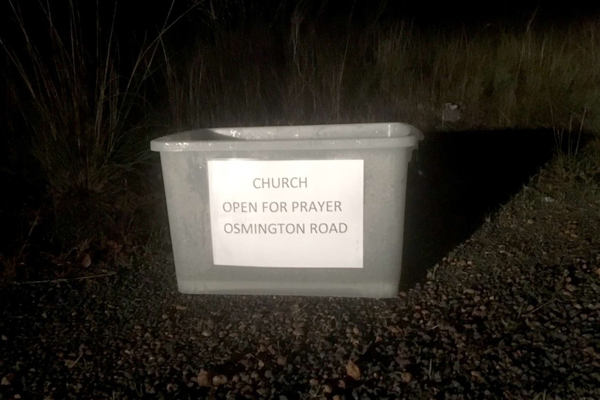 In the dark on the side of a road a white trug with a printed sign read "Church open for prayer. Osmington Road."