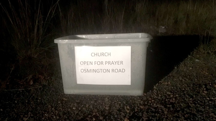 In the dark on the side of a road a white trug with a printed sign read "Church open for prayer. Osmington Road."