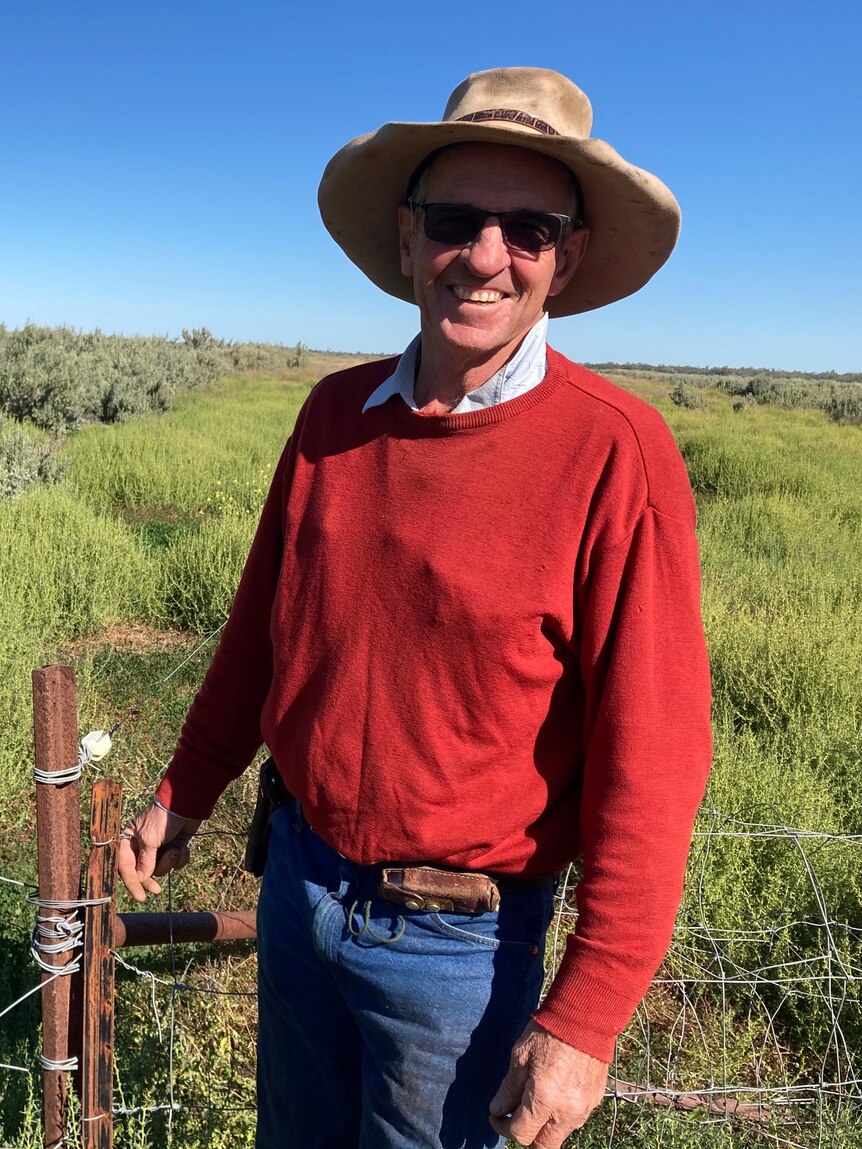 A man wearing a red jumper, jeans and a hat stands near a fence on a farm
