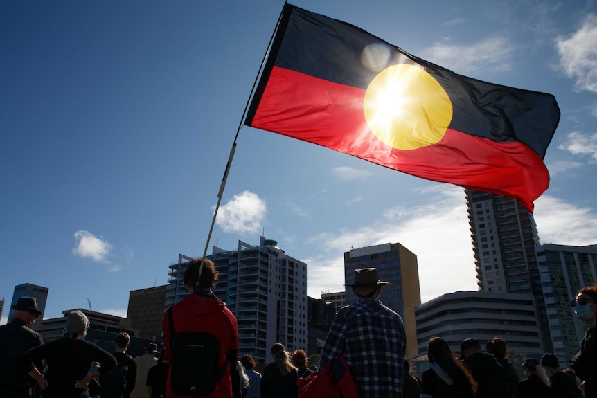Large Aboriginal flag flying in sky with sunlight behind in city setting