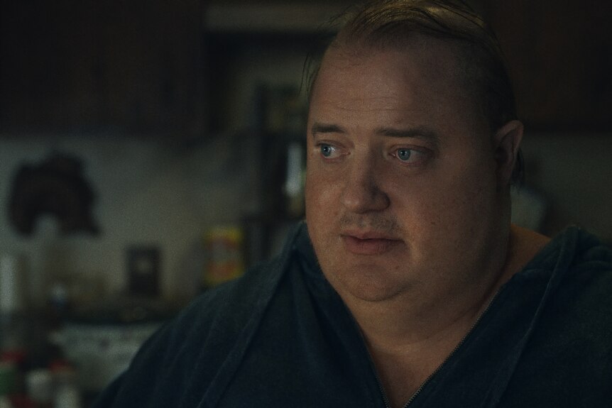 White middle-aged, overweight and balding man with blue eyes sits in a darkened room with a forlorn expression.
