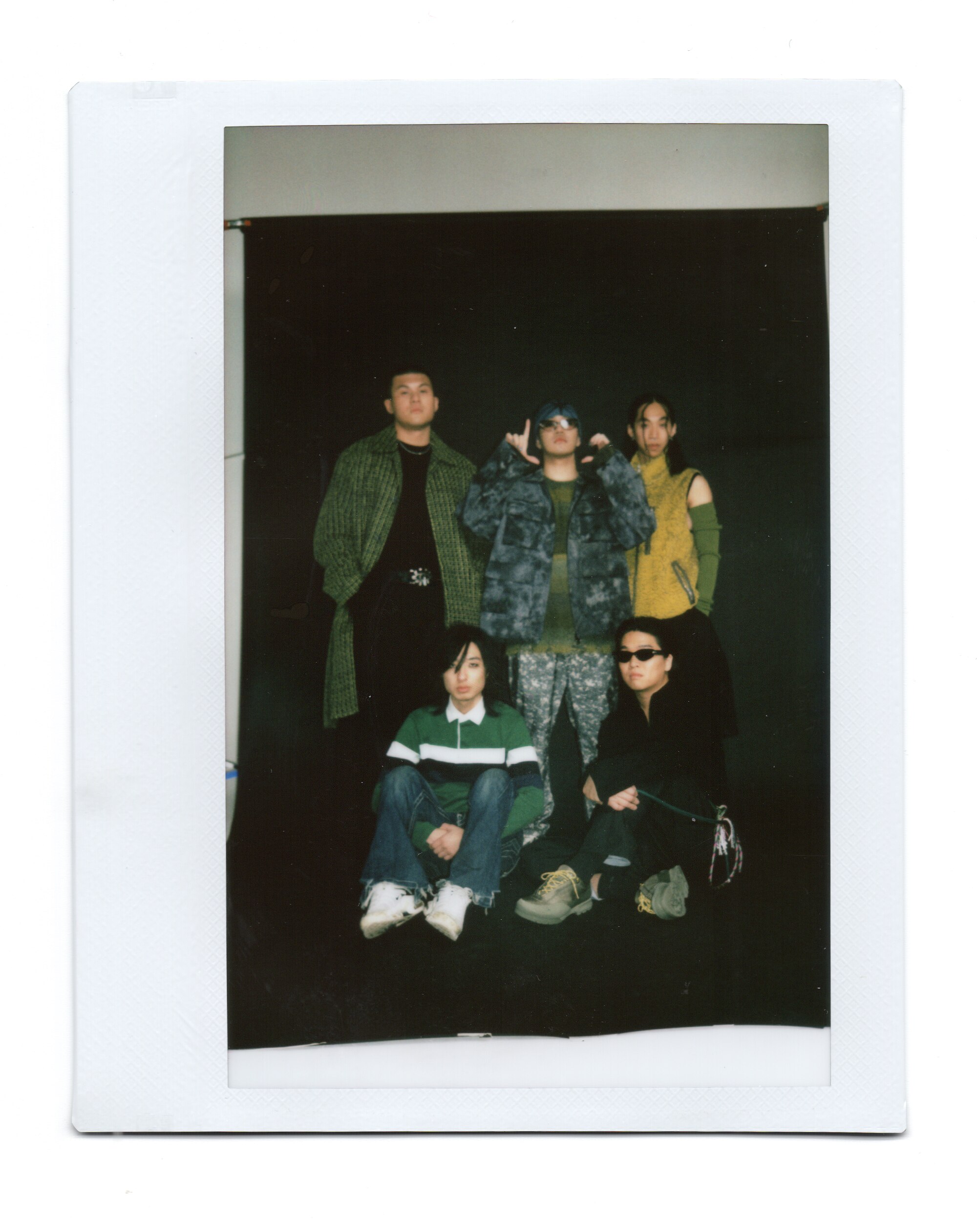 Polaroid of five men in streetwear, three are standing and two are sitting at their feet.