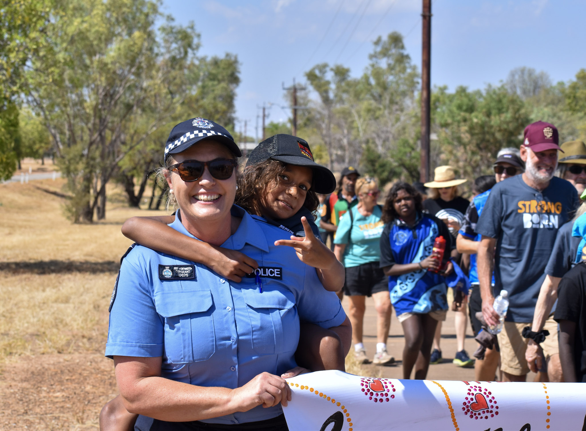 A smiling policewoman carries a young child on her back, while she holds a banner, with other marchers visible behind.