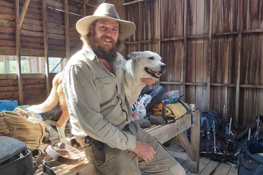 a man leans against a table with an arm around a dog surrounded by camping gear