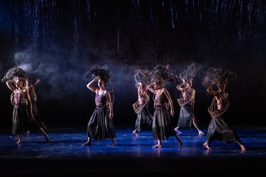 Five Indigenous dancers on a stage, wearing skirts and holding smoking feathered headdresses, the stage bathed in dark purple