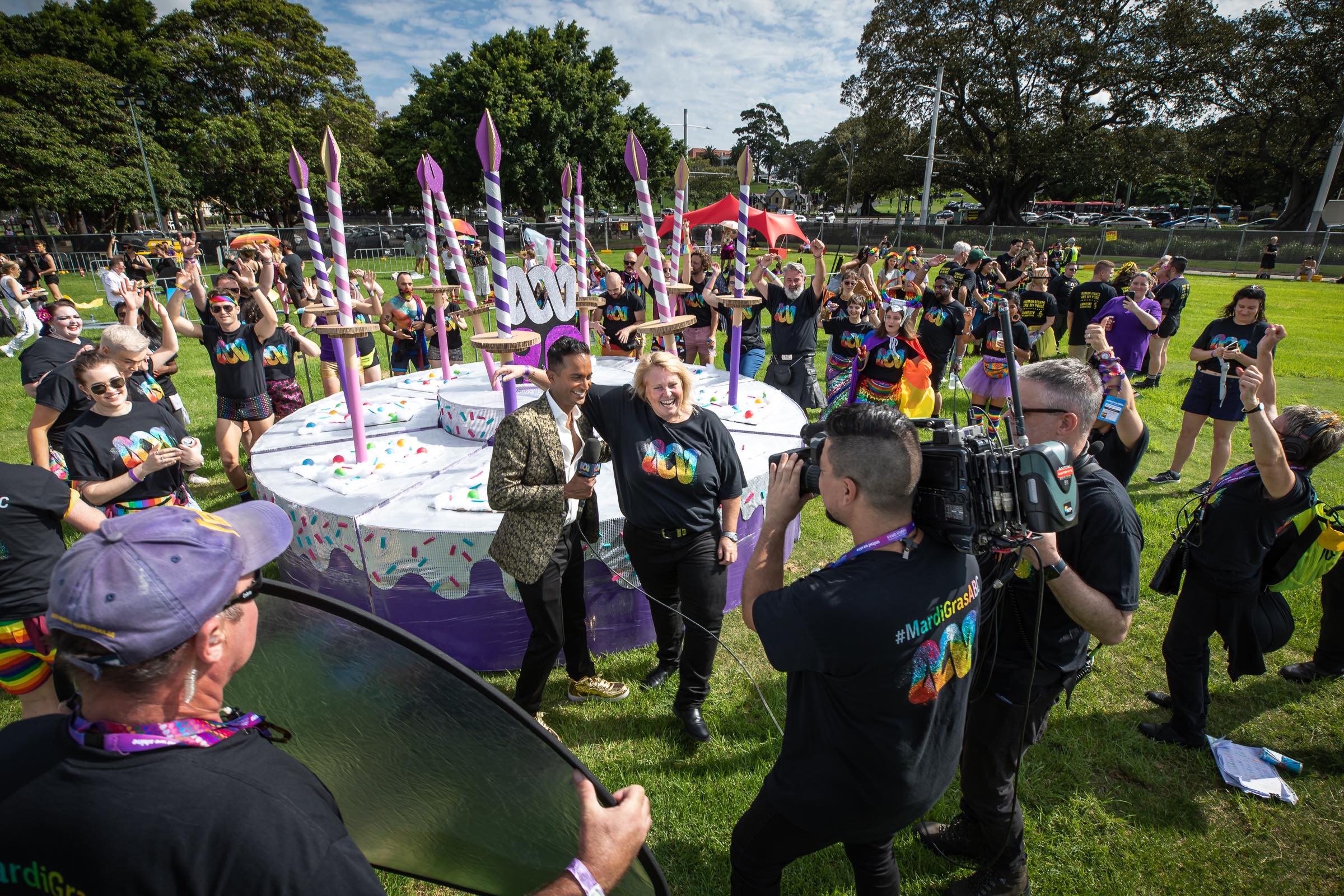 A group of happy cheering people wearing ABC Mardi Gras t-shirts surrounding a very large fake cake with candles.