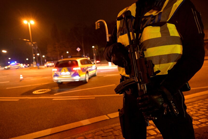 An officer holds a weapons with a police car with its lights on in the background.