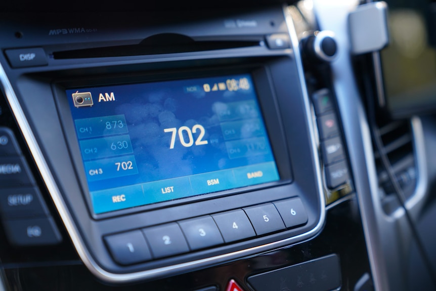 A close-up on the centre console of a car shows the radio tuned to frequency 702 AM radio.