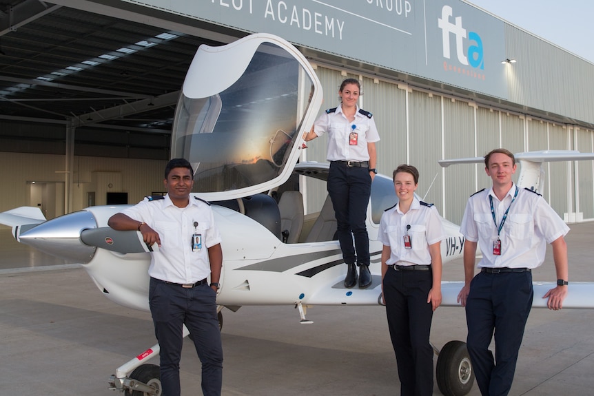 Four students in pilot uniforms stand on or next to a small single engine plane, next to a hangar. 