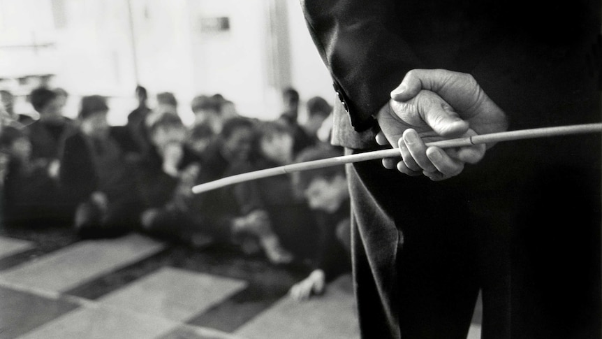 An adult holds a cane behind their back while looking out onto a classroom of students.