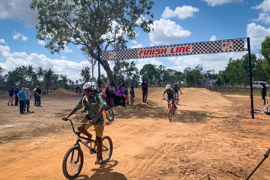 Kids race BMXs on a dirt track as spectators stand and watch.