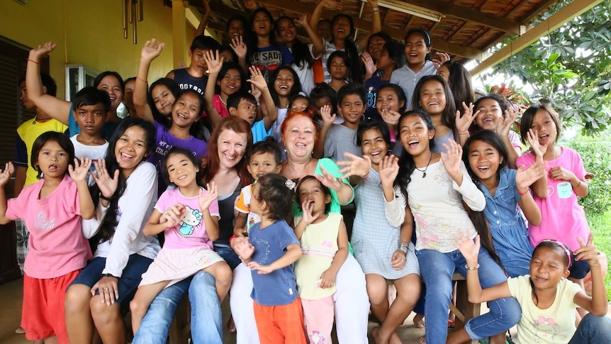 two women sitting in the middle surrounded by dozens of children smiling and waving