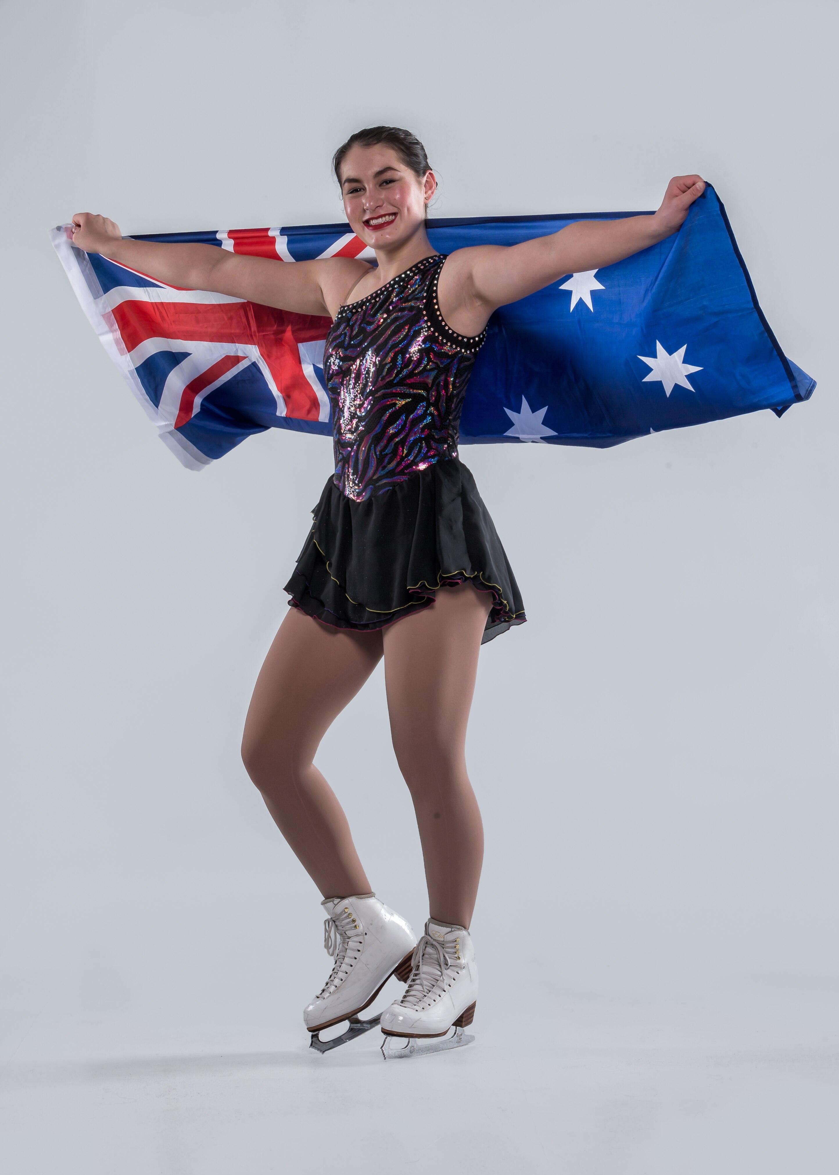 Ada Lacey, a young woman, wears ice skates and a performance leotard, while holding the Australian flag up behind her shoulders.